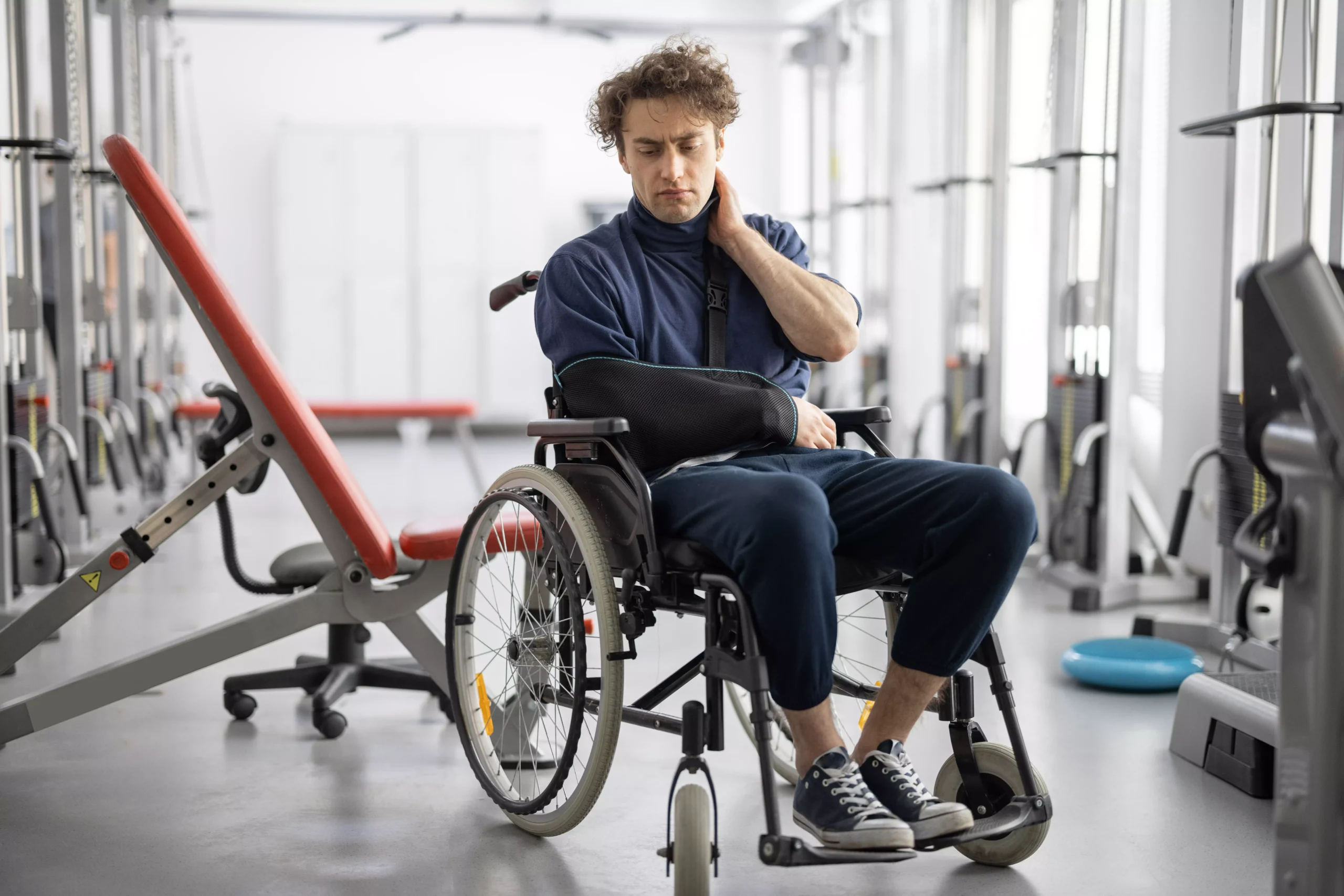 person in a wheelchair with a sling on their arm sits in a gym with exercise equipment nearby